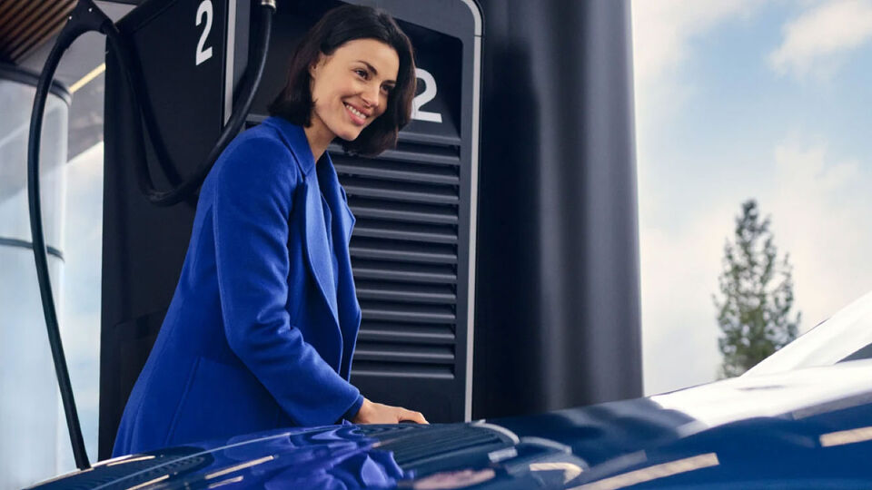 Frau steht steht neben einer Ladestation und steckt das Ladekabel an